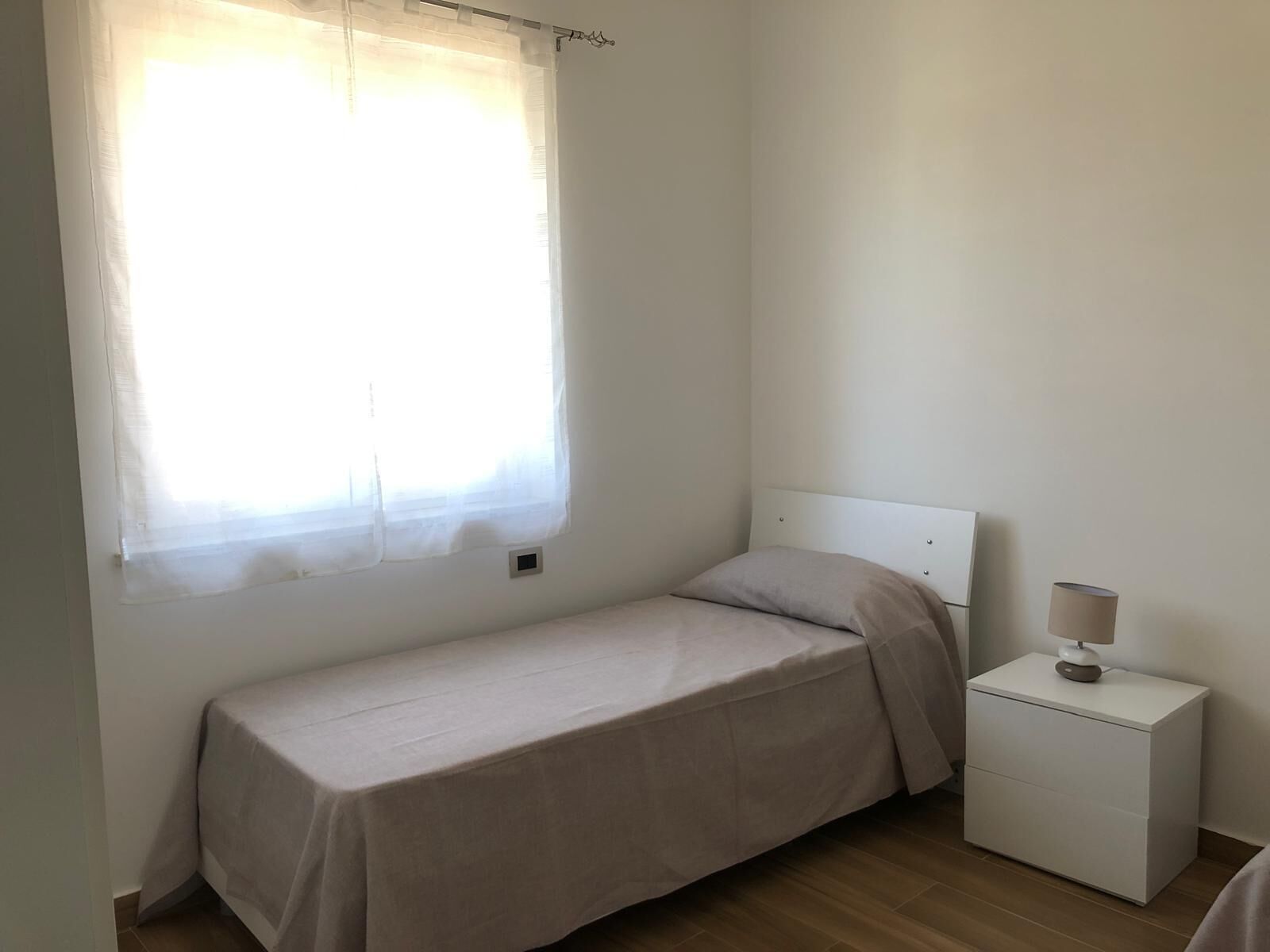 Ein kleines Zimmer mit einem Einzelbett, einem schlichten weißen Kopfstück, einem Nachttisch mit Lampe und einem Fenster mit transparenten Vorhängen.