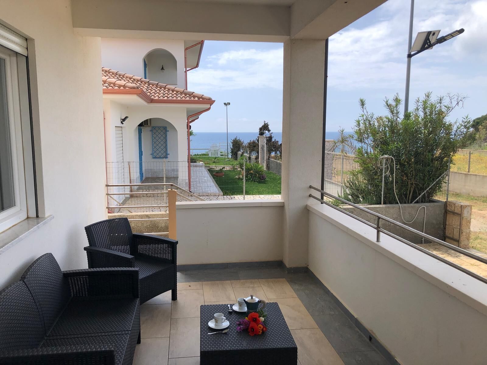 Ein Balkon mit Korbmöbeln, einem kleinen Tisch mit Tassen und Blumen sowie Blick auf ein Gebäude mit Ziegeldach und einem Tor.