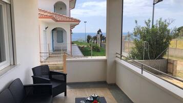 Ein Balkon mit Korbmöbeln, einem kleinen Tisch mit Tassen und Blumen sowie Blick auf ein Gebäude mit Ziegeldach und einem Tor.