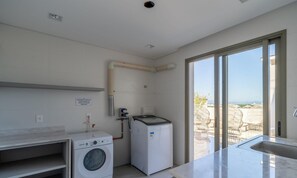 A compact kitchen with a washing machine, a small refrigerator, and a window leading to an outdoor seating area. - Ap in a Condominium With gym in Jurere Vox0210 (Florianópolis)