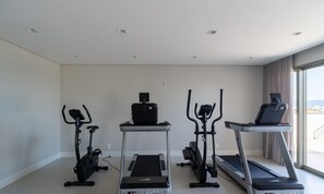 A modern hotel room with three exercise machines: a stationary bike, a treadmill, and a stair climber. The room has a large window offering a view of the cityscape. - Ap in a Condominium With gym in Jurere Vox0210 (Florianópolis)