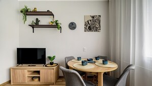 A modern dining area with a wooden table, grey upholstered chairs, a TV on a wooden cabinet, and decorative items on shelves. - RentPlanet - Apartament Zdrowa (Wrocław)