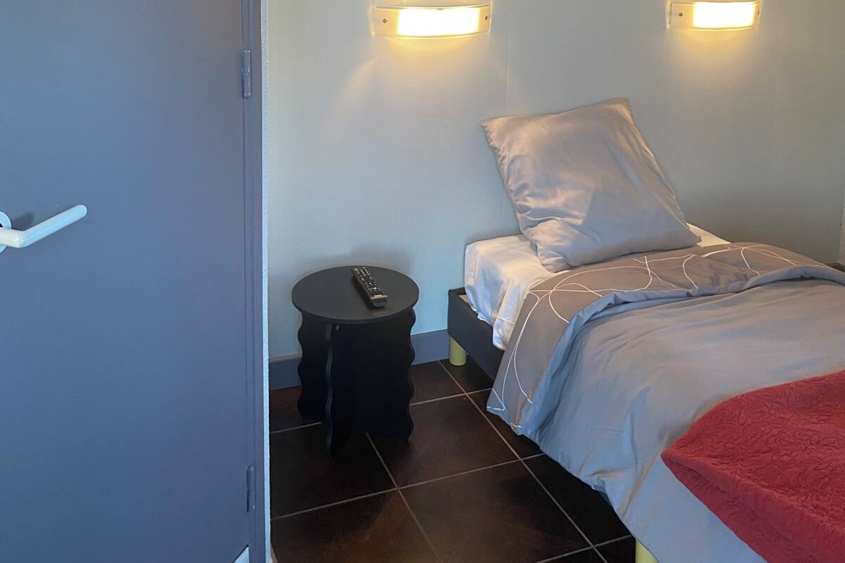A bedroom with a bed, two wall-mounted lights, a round nightstand, a remote control, and a door.