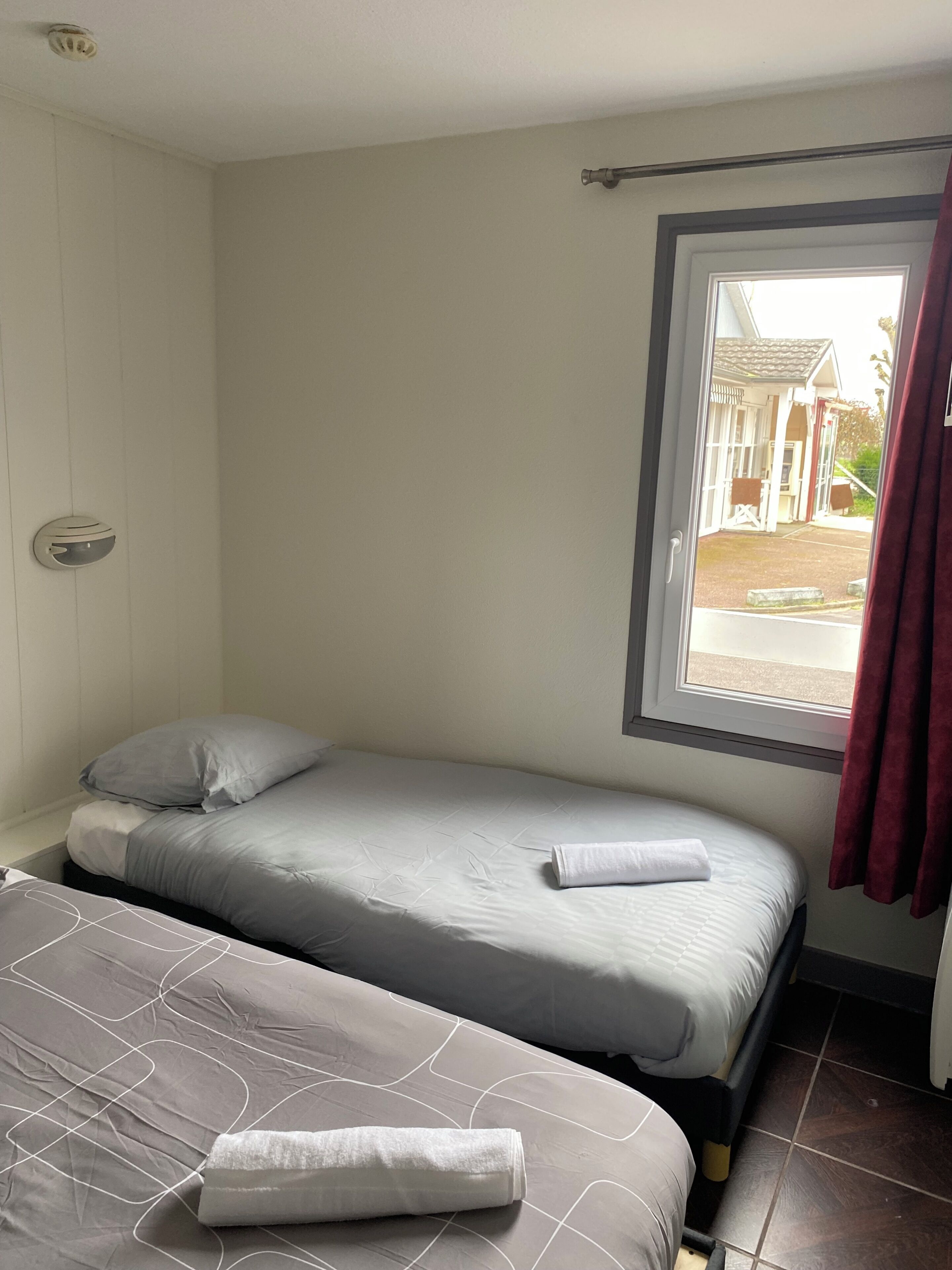 Una habitación de hotel pequeña y con una cama, dos camas, una ventana con cortinas, una puerta visible y una lámpara fijada a la pared.