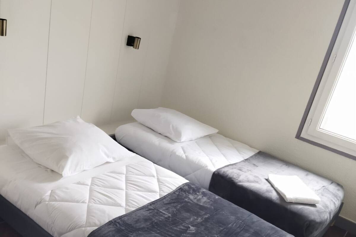 A small, compact room with two beds, a window, and a wall-mounted light fixture.