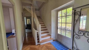 Escada que leva a um segundo andar, uma janela grande com vista para vegetação, e um tapete listrado azul na porta.