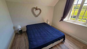 A bedroom with a bed, a nightstand, a lamp, a heart-shaped decoration on the wall, and a window with a view of greenery. - Charming cottage Les Prés (Saint-Cyprien)