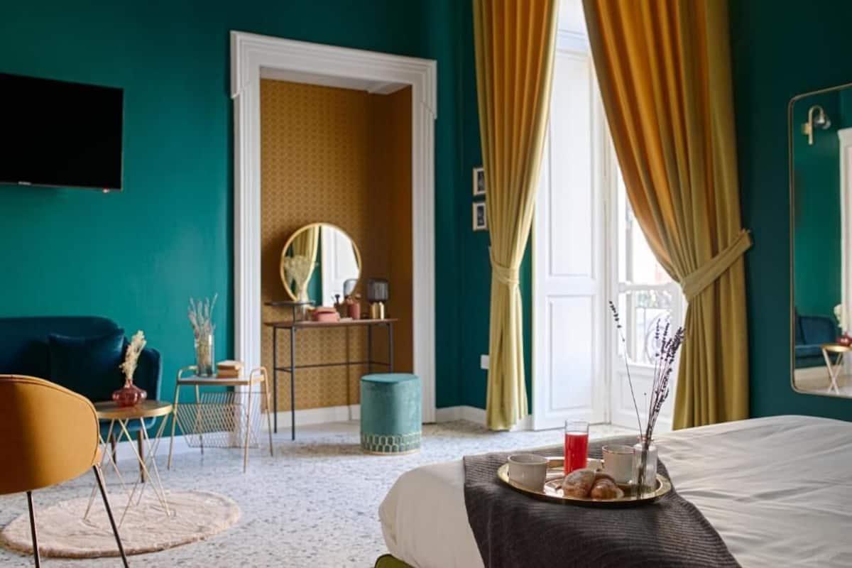 Hotellihuone, jonka seinät ovat maalattu turkoosilla, valkoisella peitteellä varustettu sänky, aamiaistarjotin, jossa on croissantteja, pieni pyöreä pöytä, jossa on maljakko, ja peili seinällä.