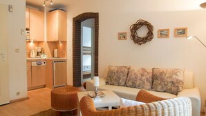 A cozy living room with a sofa, two wicker chairs, a small table, and a kitchen area with wooden cabinets and a brick archway. - Apartment 2 - Holiday home Sternmöwe Cuxhaven (Cuxhaven)
