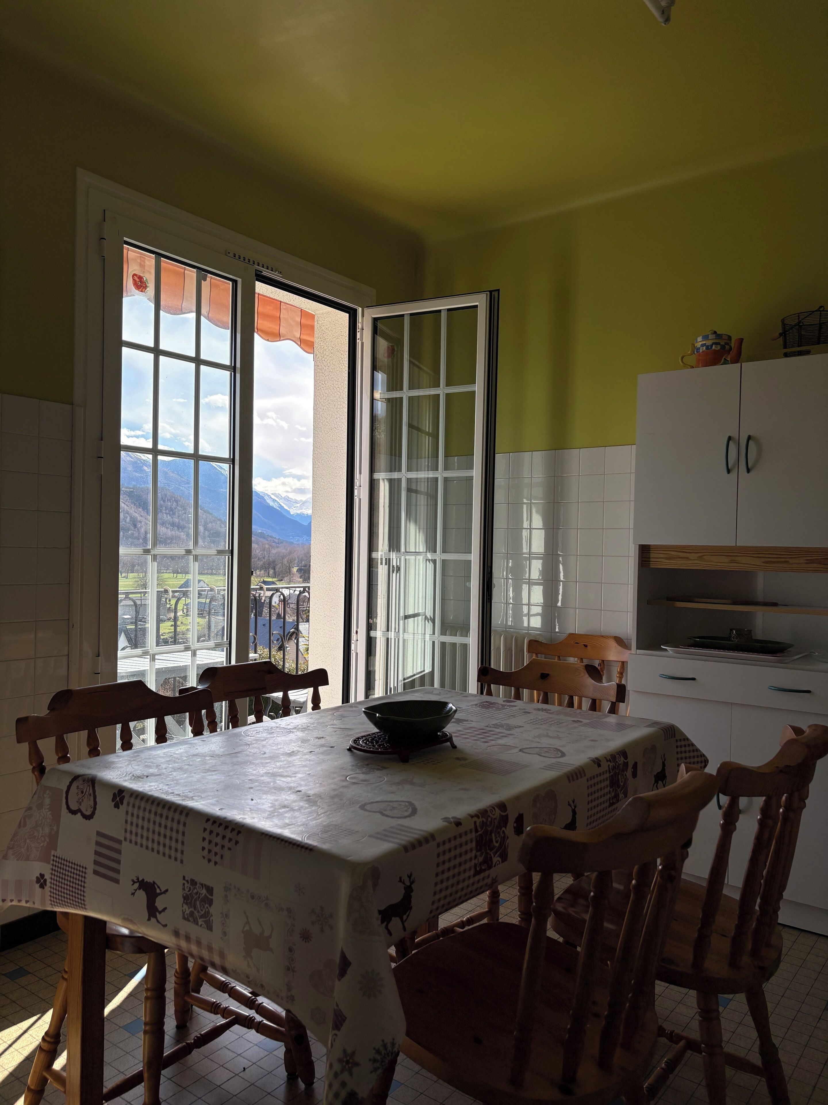 Un comedor con una mesa dispuesta para cuatro, una ventana con vistas a las montañas y una cocina con armarios blancos y una silla de madera.