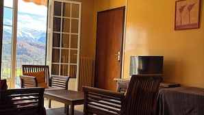 Ein Zimmer mit Holztisch und Stühlen, einem Bett mit gestreifter Decke, einem Fernseher und einer Tür zu einem Balkon mit Bergblick.