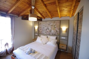 A bedroom with a wooden ceiling, a bed with white bedding, a wall-mounted light, and a floral painting. - Hameau du jardin Fleuri near Bergerac - 10 gîtes, sleeps 40 (Bosset)