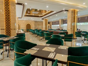 A spacious dining area with green upholstered chairs and tables, a central bar area, and a high ceiling with recessed lighting. - Grand Maya by ARTOTEL - Banjarbaru (Banjarbaru)