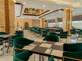 Un comedor amplio con sillas tapizadas en verde y mesas, una zona de bar central y un techo alto con iluminación empotrada.