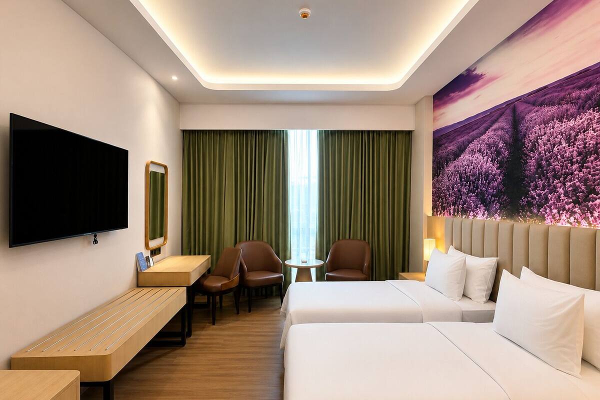 Habitación de hotel con televisión de pantalla plana, un banco de madera, dos camas individuales, una mesita pequeña, dos sillas y un amplio ventanal con cortinas verdes.