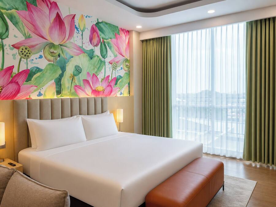Una habitación de hotel con una cama grande, un cabecero, una mesita de noche con lámpara, un banco y una ventana con cortinas.
