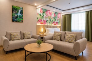 A hotel room with two sofas, a round coffee table, a painting of a waterfall, and a floral mural. - Grand Maya by ARTOTEL - Banjarbaru (Banjarbaru)