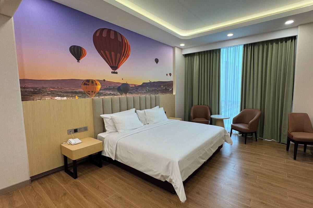 Una habitación de hotel con una cama grande, un escritorio, dos sillas, una mesita, una mesita de noche y un mural de globos aerostáticos.