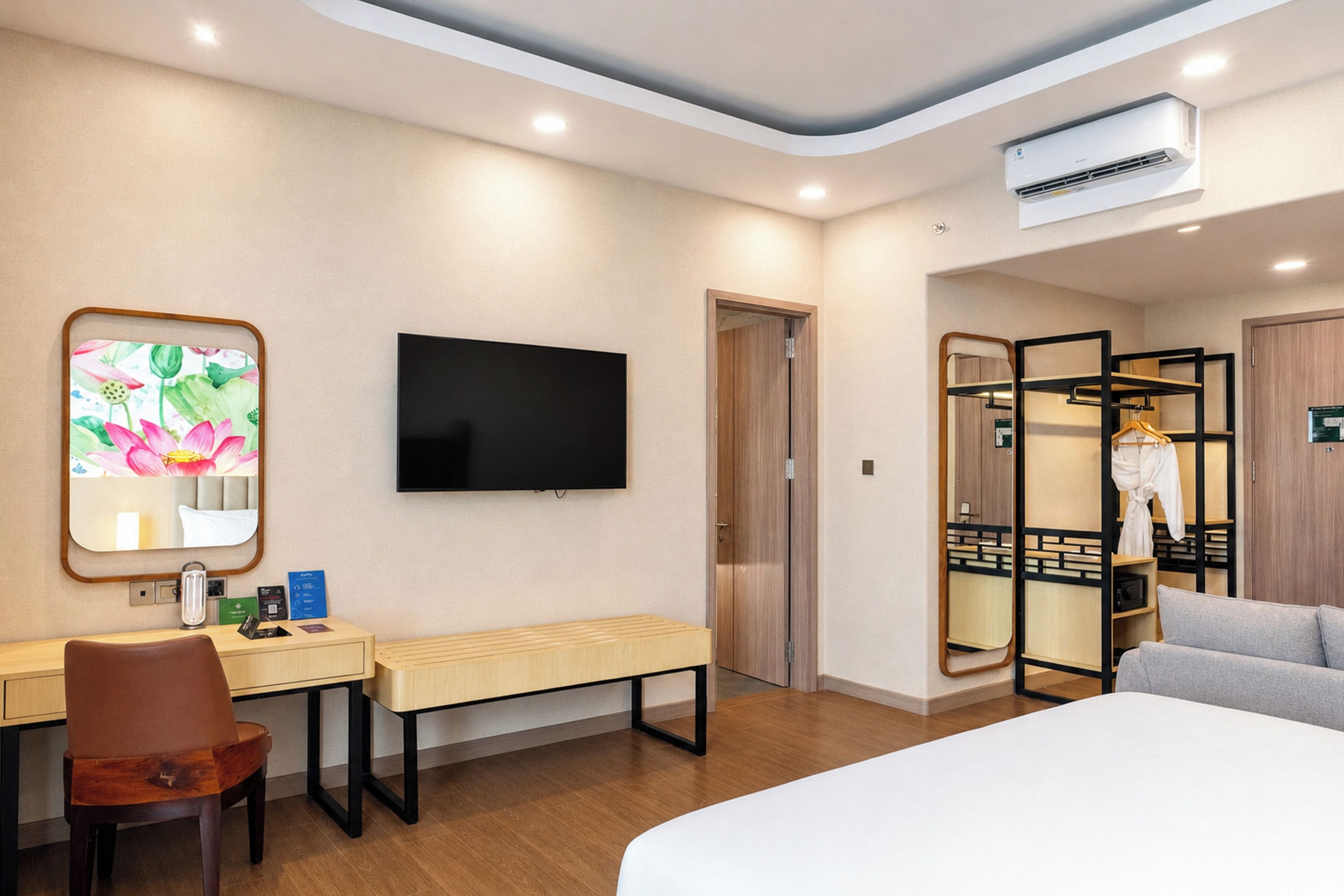 A hotel room with a bed, a desk with a chair, a television, a bench, a wardrobe, and a mirror.