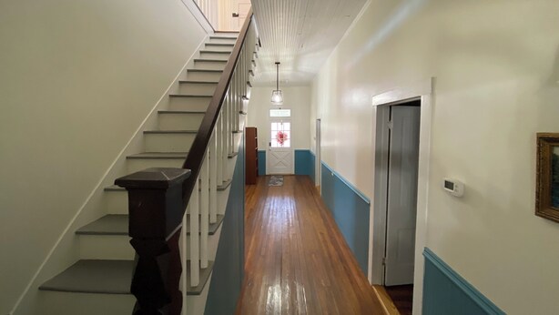 Un pasillo con piso de madera, paredes blancas y una escalera con barandillas oscuras.