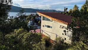 Una casa moderna con tetto in tegole rosse, facciata in pietra e vista su un lago e montagne sullo sfondo.