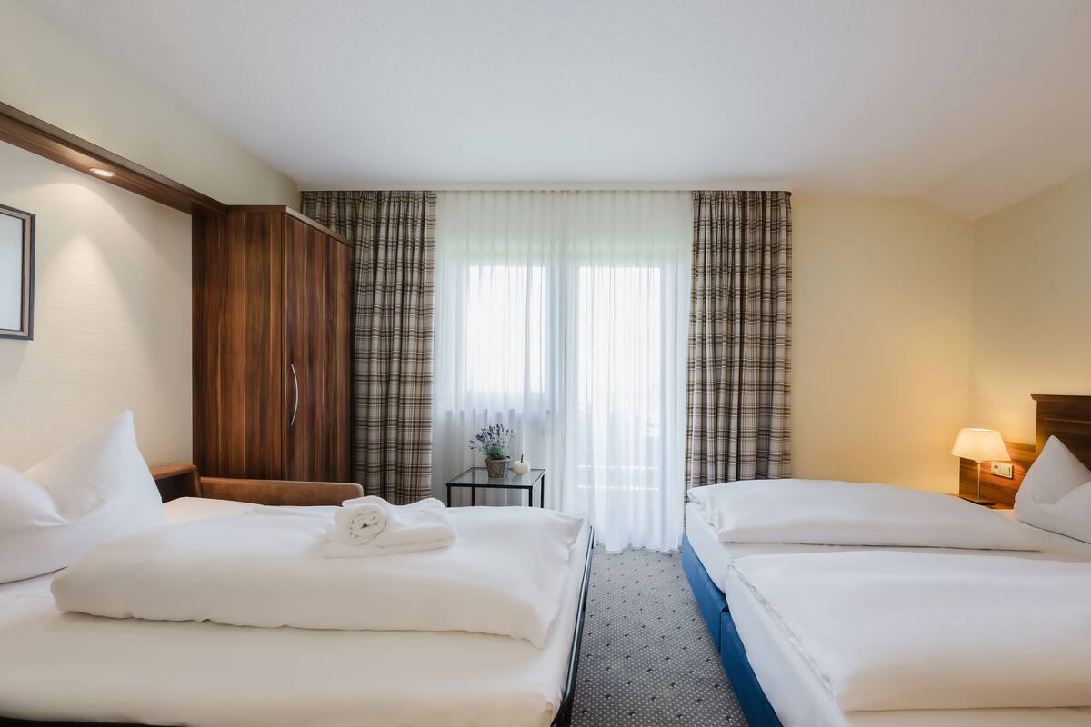 Une chambre d’hôtel avec deux lits, une armoire, une petite table avec une lampe et une vue dégagée à travers des rideaux légers.