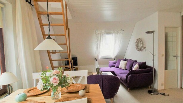 Uma sala de estar aconchegante com uma escada de madeira, um sofá roxo, uma mesa branca com um vaso de flores e um abajur.