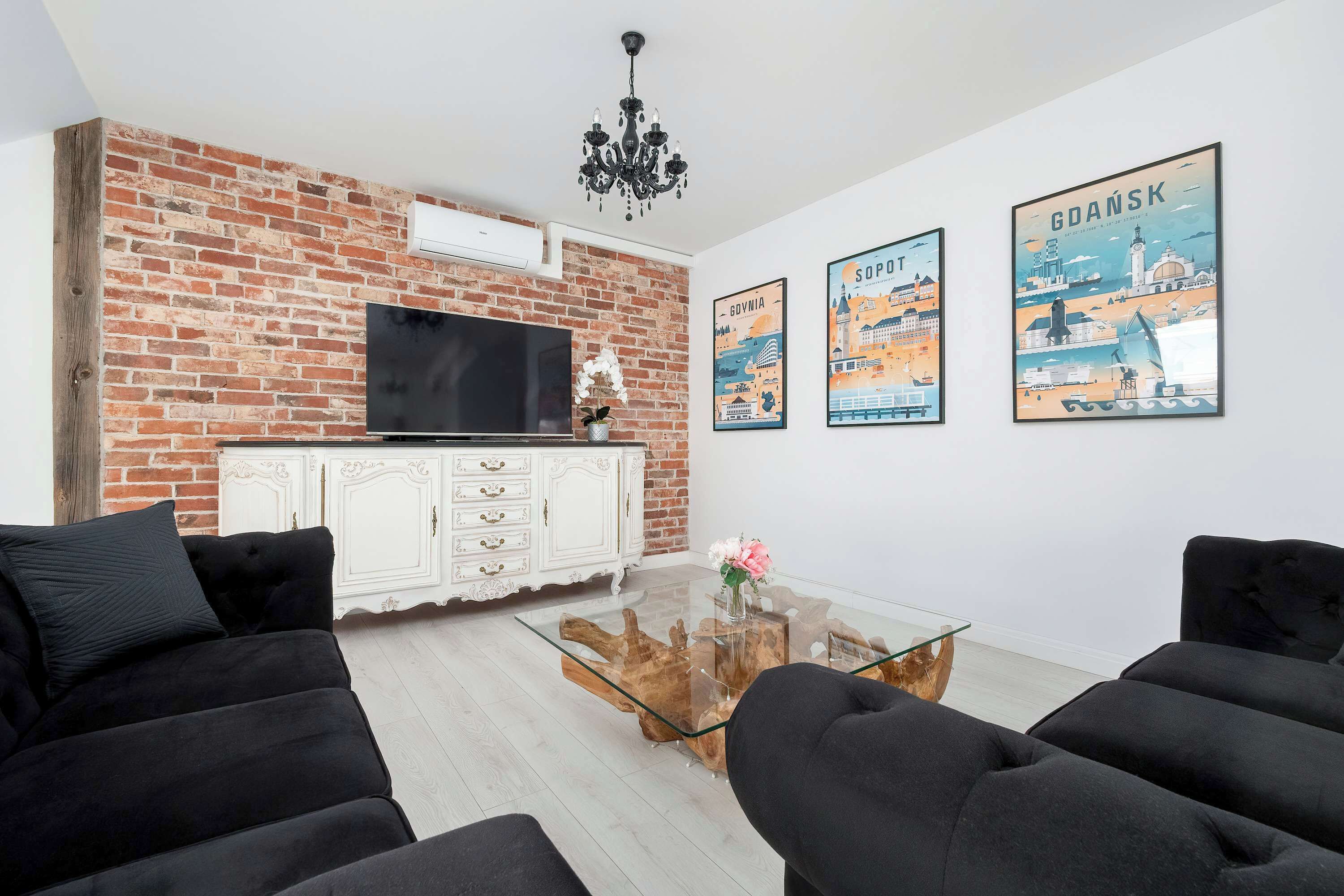 Ein Wohnzimmer mit Backsteinwand, einem Flachbildfernseher, einem weißen Schrank, schwarzen Sofas und drei Reiseplakaten.
