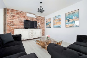 Ein Wohnzimmer mit Backsteinwand, einem Flachbildfernseher, einem weißen Schrank, schwarzen Sofas und drei Reiseplakaten.