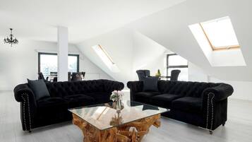 Ein modernes Wohnzimmer mit zwei schwarzen Sofas, einem Glastisch und einem Couchtisch mit Holzboden. Es gibt Dachfenster und eine Kristallleuchter.