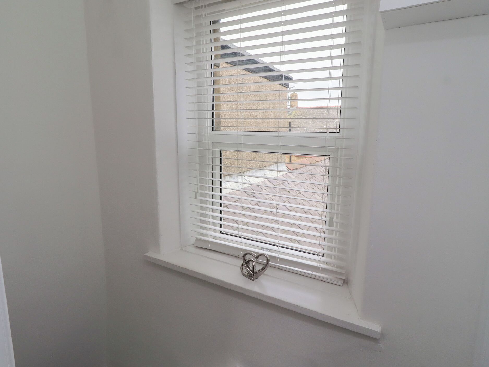 Une fenêtre avec des stores blancs, un décor en forme de cœur sur le rebord, et une vue sur un bâtiment à l’extérieur.
