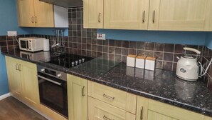A kitchen with light wood cabinets, a black countertop, a microwave, a kettle, and a stove. - 23 Northumbria Terrace (Morpeth)