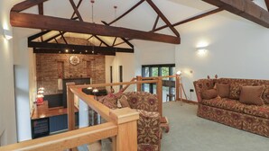 A spacious living area with a high ceiling and exposed wooden beams, a cozy patterned sofa, and a stone wall with a clock. - The Barn at Sunny Barn (Brecon)