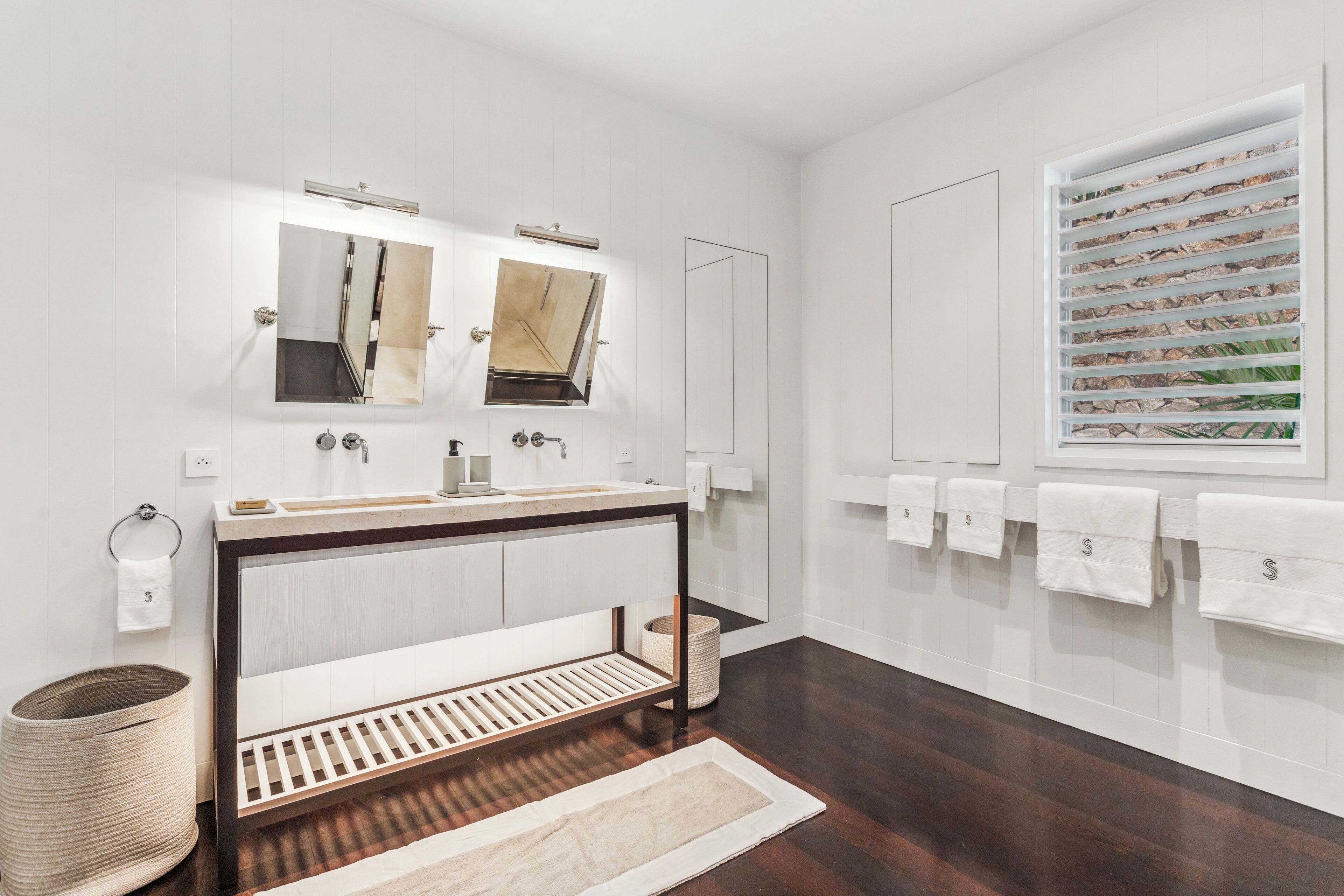 A modern bathroom with a large mirror, dual sinks, and a window with a view of plants.