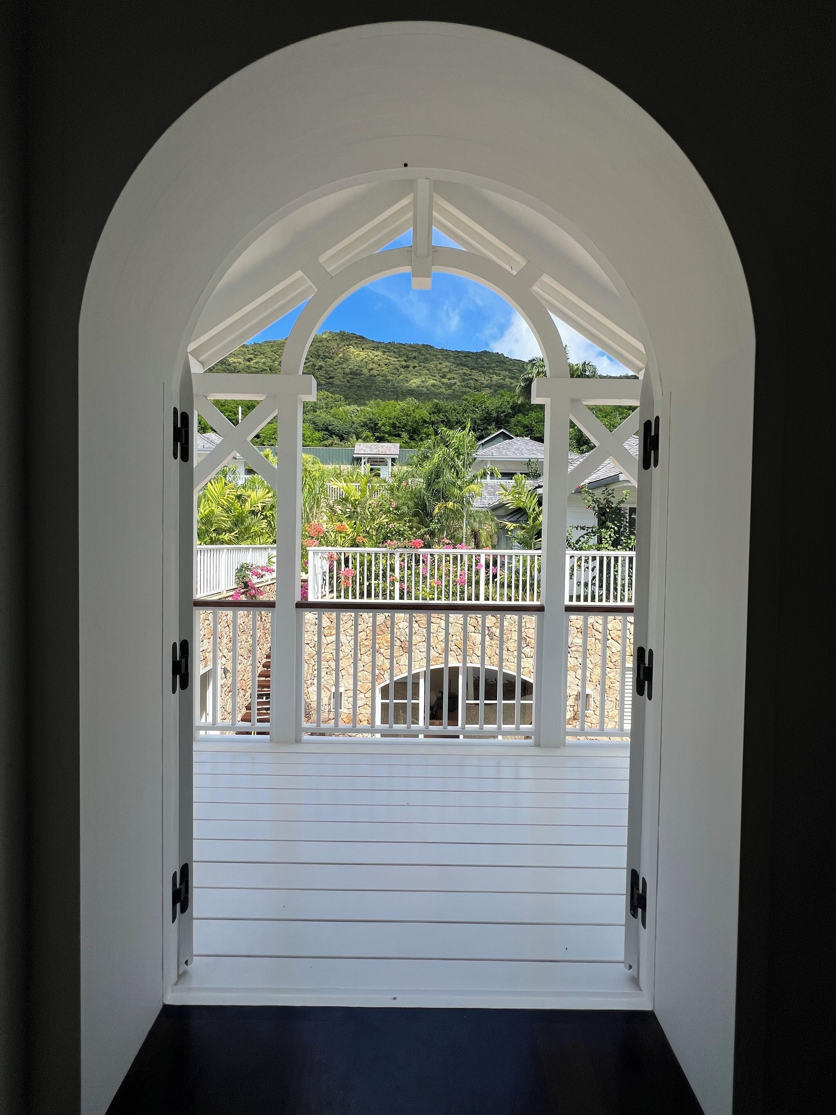 Un arco de entrada conduce a un balcón con un muro y barandilla de piedra, con vistas a un paisaje verde exuberante y casas.