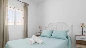 Una cama bien tendida con cubrecama acolchado, una mesita de noche con lámpara y una ventana con cortinas.