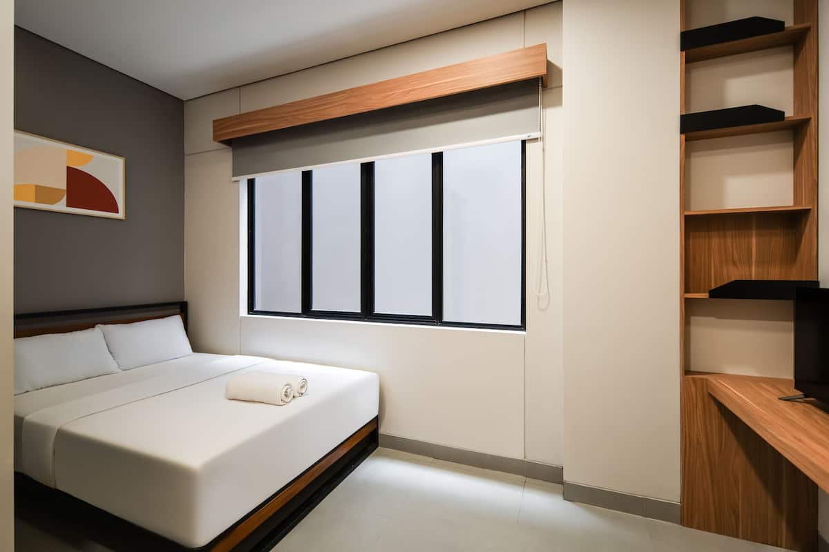 A modern bedroom with a bed, a window with blinds, a wooden shelf unit, and a framed abstract artwork on the wall.