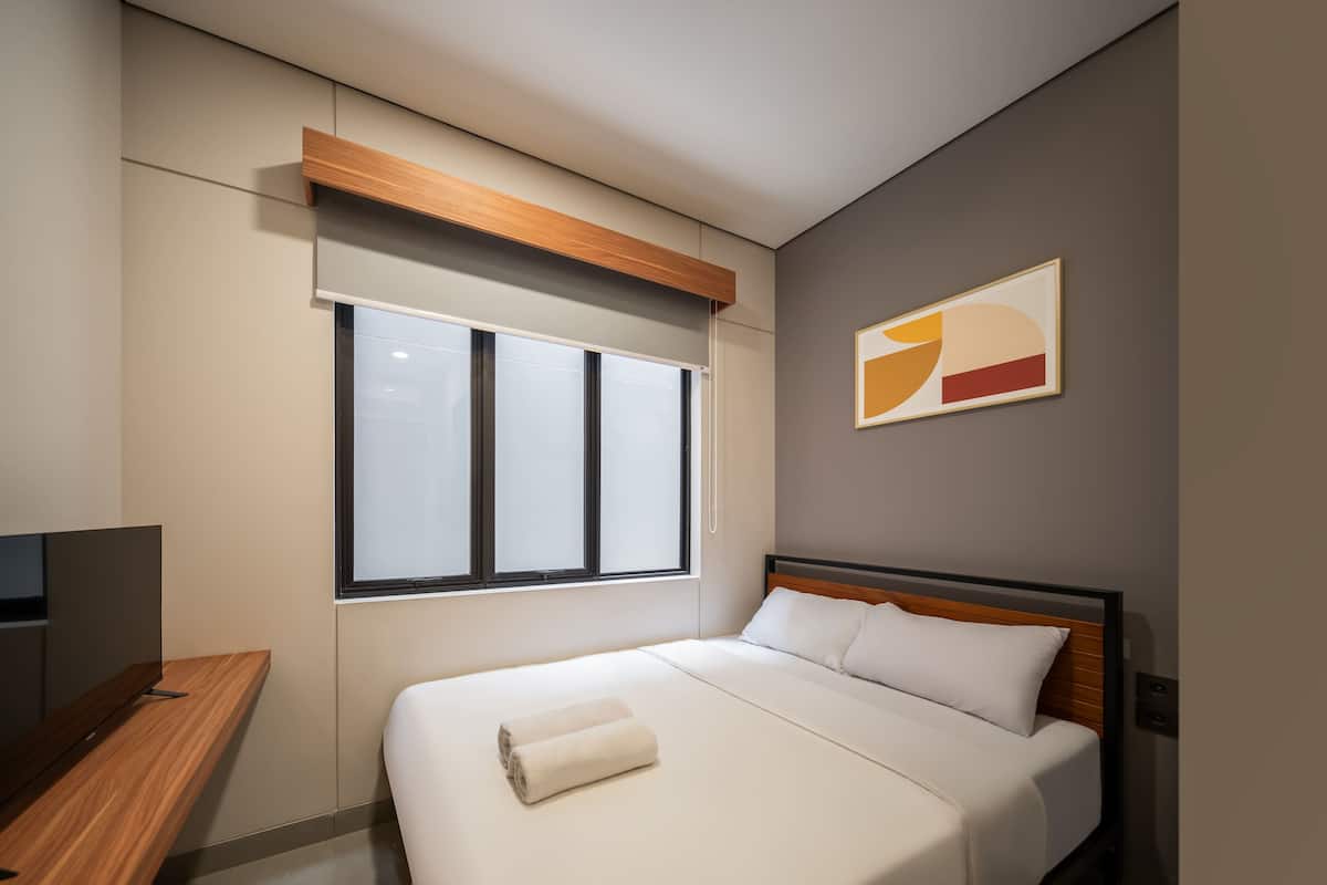A modern hotel room with a bed, a wooden bedside table, a window with blinds, and a wall-mounted abstract art piece.