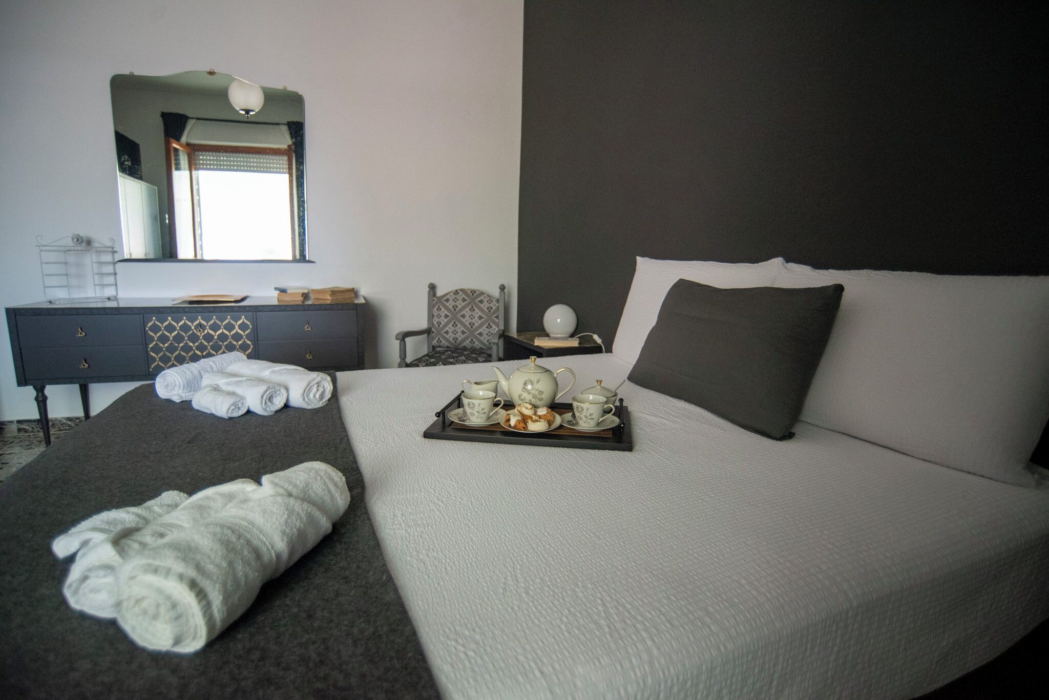 A neatly made bed with a tray of tea set, towels, and a chair with a patterned backrest.
