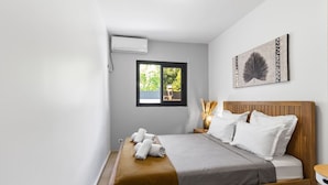 A bedroom with a wooden bed, white pillows, a gray blanket, a window with a view of greenery, and a wall-mounted air conditioner.