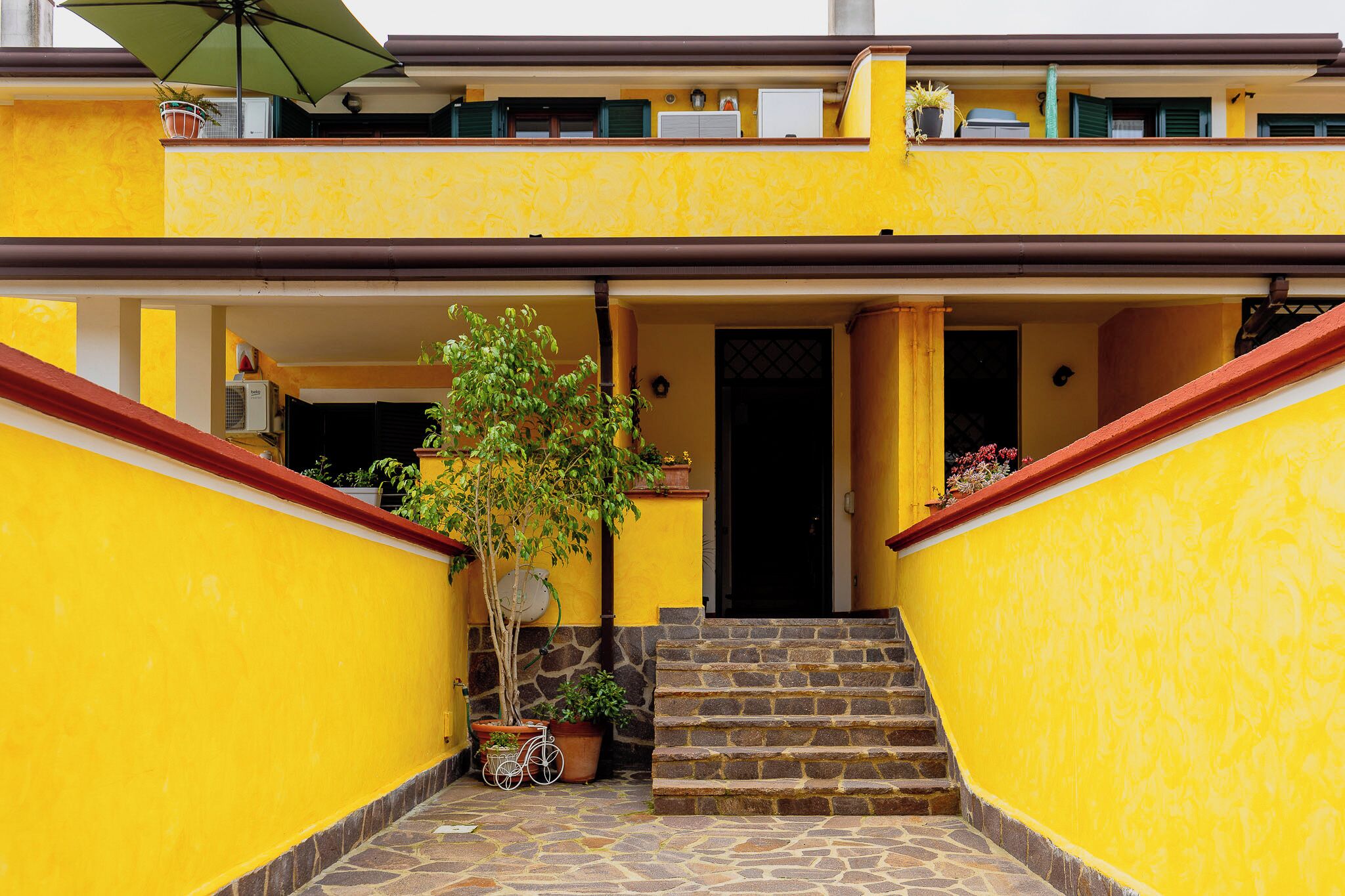 Un bâtiment jaune avec un escalier menant à une porte, des plantes en pot et un vélo.