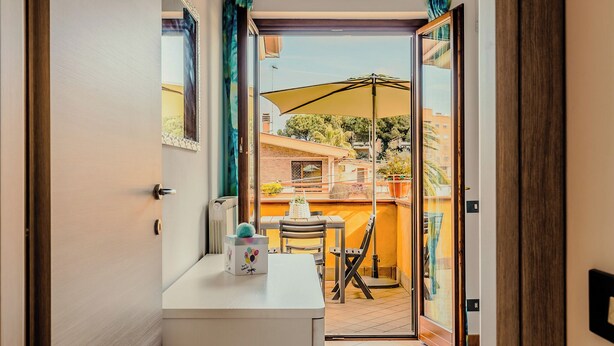 A room with a sliding glass door leading to a balcony with a table and chairs, a large umbrella, and a view of buildings and trees.