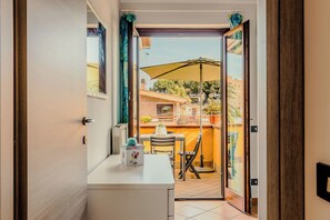 Een kamer met een schuifdeur naar een balkon, met een tafel, stoelen, een grote parasol en uitzicht op gebouwen en bomen.