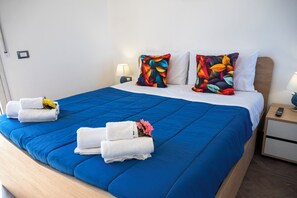 A neatly made bed with blue bedding, white pillows, and colorful floral pillows. There are towels and a small vase with flowers on the bed. - Villa Verde- Nature Holiday Home (Carini)