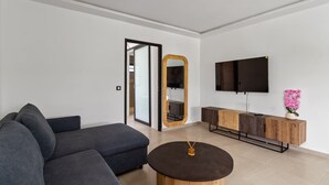Uma sala de estar moderna com um sofá modular cinza, uma mesa de centro redonda, um armário de madeira, uma televisão de tela plana e um espelho.