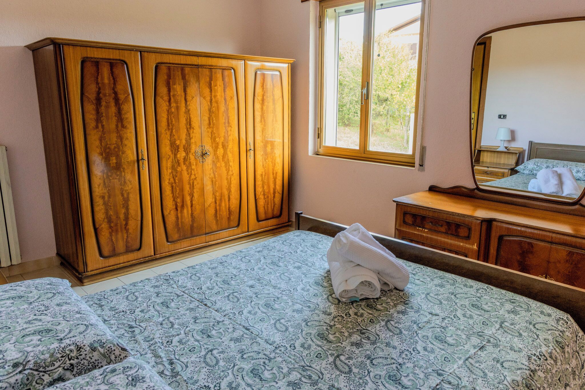 A bedroom with a large wooden wardrobe, a bed with a patterned bedspread, a window with a view of greenery, and a bedside table with a lamp.