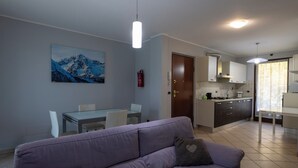 A modern living room with a purple sofa, a dining table with chairs, a kitchen area with cabinets and a stove, and a painting of mountains on the wall. - La Maison De L'Adret (Saint-Pierre)