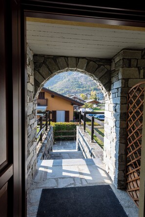 Un arc en pierre mène à un bâtiment doté d’un balcon, d’une clôture en bois et offrant une vue sur une montagne.