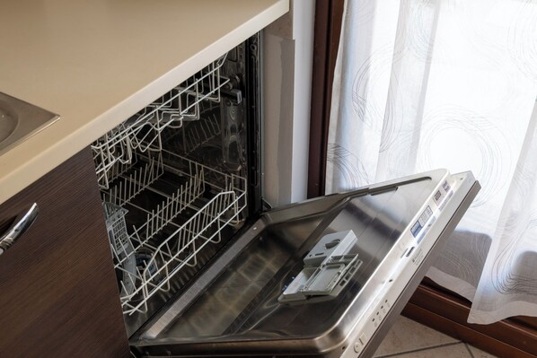 An open dishwasher with a metal rack inside, a control panel on the right, and a kitchen counter to the left. - La Maison De L'Adret (Saint-Pierre)