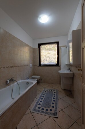 Une salle de bain avec une baignoire, des toilettes et un lavabo. Il y a une fenêtre donnant sur un paysage verdoyant.